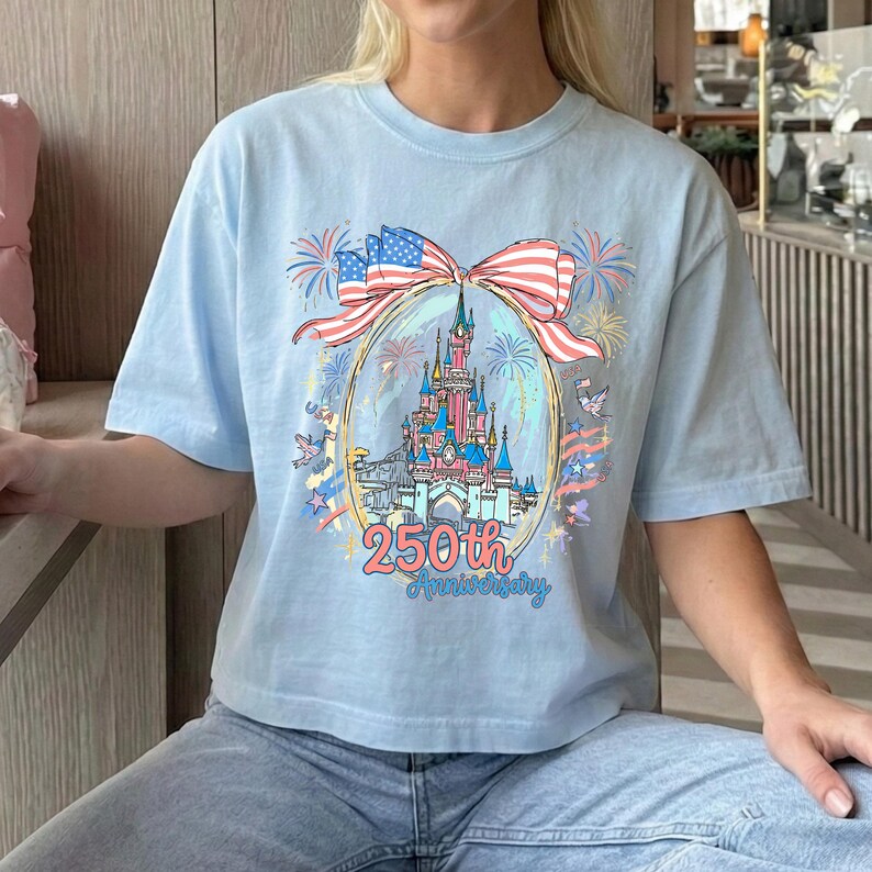 K&ouml;nnte beinhalten: Hellblaues T-Shirt mit einem farbenfrohen Aufdruck eines Schlosses, Feuerwerks und einer Schleife im Stil der amerikanischen Flagge. Der Text "250th Anniversary" ist darunter. Das T-Shirt hat einen l&auml;ssigen Stil.