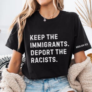 May include: Black t-shirt with the text "KEEP THE IMMIGRANTS. DEPORT THE RACISTS." in white block letters. The sleeve reads "ABOLISH ICE" in white. The shirt is worn by a person.