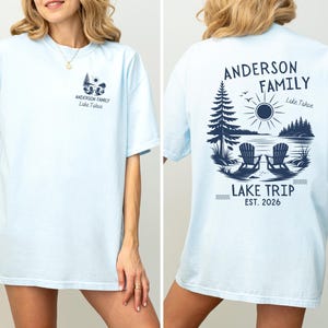 May include: A light blue short-sleeved t-shirt featuring a dark blue graphic print. The front displays a small lake scene with two chairs and the text "Anderson Family Lake Tahoe." The back features a larger version of the same graphic.