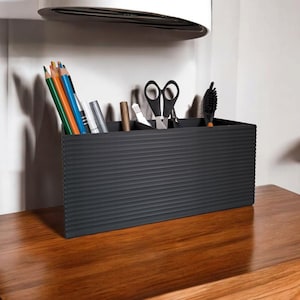 May include: A black desk tidy with a ribbed design, containing stationery items. The organiser holds coloured pencils, pens, scissors, and a small brush. It is placed on a wooden surface.