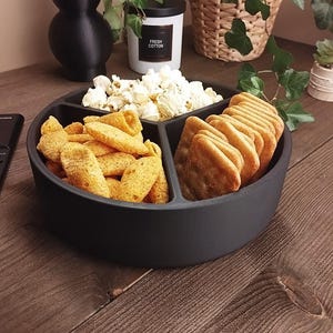 May include: A black serving platter divided into sections, filled with snacks. It holds popcorn, crackers, and crisps. The platter is on a wooden surface, with a candle and plants in the background.
