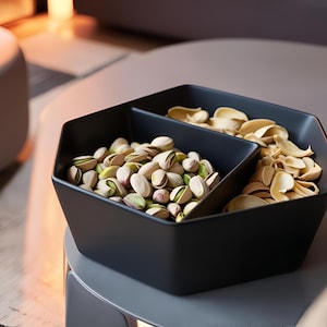 May include: A black, geometric serving bowl filled with pistachios and shells. The bowl is divided into two sections, one holding whole pistachios and the other shells. The pistachios are green and tan.
