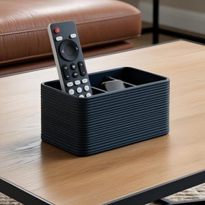 May include: A dark blue, ribbed remote control tidy on a wooden table. A white and black remote control is in the tidy, along with a small, grey device. The tidy has multiple compartments.