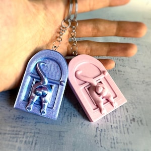May include: Two keychains with a cat design. One is a gradient of blue and purple, the other is pink. Each keychain features a cat's rear end and tail, with a door frame design. The keychains are attached to silver keyrings.