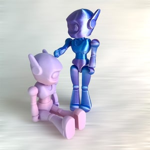 May include: Two articulated robot figures in shades of purple and blue. One figure is seated, the other standing, with one arm extended. The figures have rounded heads, ear-like protrusions, and jointed limbs, suggesting a futuristic or sci-fi theme.