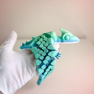 May include: A 3D-printed, stylized teal and blue dinosaur figurine. The figurine has a segmented body with a textured pattern, a pointed head, and small arms. The color transitions from teal to blue.