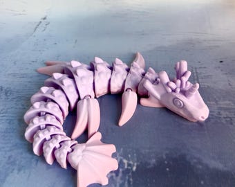 Könnte beinhalten: Ein bewegliches Drachenspielzeug in Lavendel mit einem segmentierten Körper, Flossen und einem detaillierten Kopf. Der Drache besteht aus Kunststoff und hat eine glatte, matte Oberfläche. Das Spielzeug ist beweglich und flexibel, wobei sich jedes Segment unabhängig bewegen kann.