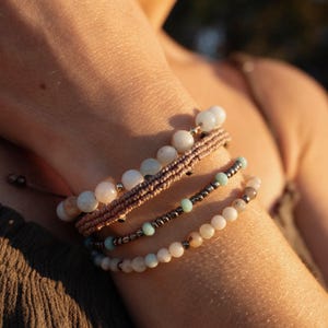 Può includere: Primo piano di un polso che indossa quattro braccialetti. I braccialetti sono realizzati con materiali diversi, tra cui perline, corda e metallo. Le perline sono di vari colori, tra cui bianco, azzurro e marrone.