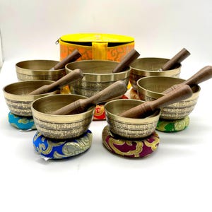 Könnte beinhalten: Satz von sieben Klangschalen mit Holzschlägeln und bunten Kissen. Die Schalen sind aus Metall und haben komplizierte Designs. Die Schalen sind in einem Halbkreis angeordnet, wobei sich die Hülle im Hintergrund befindet.