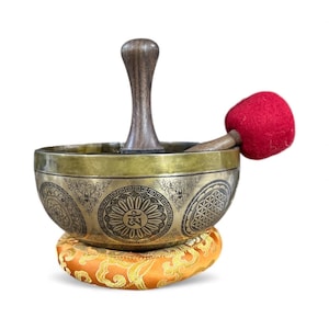 May include: A brass singing bowl with intricate etched patterns, resting on an orange cushion. A wooden mallet with a red felt head rests beside the bowl. A wooden handle is attached to the bowl.