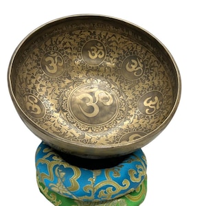 Może przedstawiać: Brązowa miska śpiewająca z symbolami Om i skomplikowanymi wzorami. Misa spoczywa na niebiesko-zielonej poduszce we wzory. Symbol Om jest wyraźnie widoczny pośrodku.