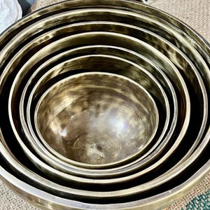 Puede incluir: Una colección de cuencos cantores de color bronce de varios tamaños, anidados unos dentro de otros. Los cuencos tienen un acabado metálico liso y están dispuestos en un patrón circular. La imagen está tomada desde un ángulo ligeramente elevado, mostrando la profundidad y forma de los cuencos.