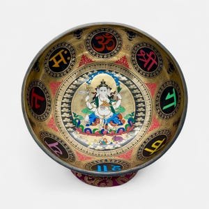 Pode incluir: Uma tigela cantora dourada com desenhos intrincados. A tigela apresenta uma imagem central de uma divindade, rodeada por símbolos e padrões coloridos. Descansa sobre uma base com padrões roxos e dourados.