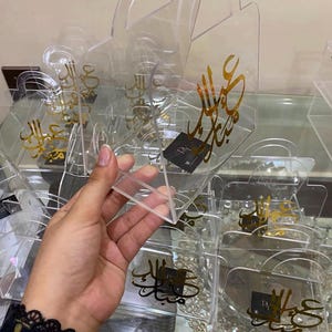 Può includere: Sacchetti regalo in acrilico trasparente con calligrafia araba dorata. Le borse hanno un manico e una base rettangolare. Il testo sulle borse dice "Eid Mubarak". Un piccolo quadrato nero con una "R" bianca è sulle borse.