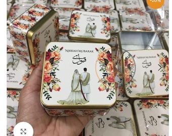 以下が含まれることがあります： 花柄と金色の縁取りが施された正方形の金属製の缶。蓋にはウェディングドレスを着たカップルと「Nikkah Mubarak」の文字が描かれています。これらの缶は、結婚式の引き出物やギフトとして使用される可能性があります。