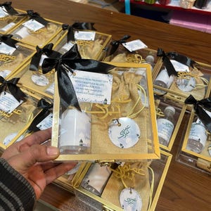 Peut inclure: Plusieurs coffrets cadeaux avec des cadres dorés, des nœuds noirs et divers contenus. Chaque boîte contient une petite bouteille, une pochette et une étiquette décorative. Le texte sur l'étiquette indique "Blessings from Prophet City with gratitude & lots of duas for you."