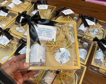 May include: Several gift boxes with gold-colored frames, black bows, and various contents. Each box contains a small bottle, a pouch, and a decorative tag. The text on the tag reads "Blessings from Prophet City with gratitude & lots of duas for you."