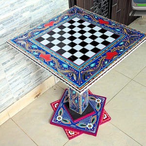 Puede incluir: Mesa de ajedrez cuadrada con tablero a cuadros blanco y negro. La mesa presenta un diseño pintado a mano con patrones florales azules, rojos y dorados en los bordes. Se apoya en una base de pedestal decorativa. Dimensiones: aprox. 76 cm x 76 cm.