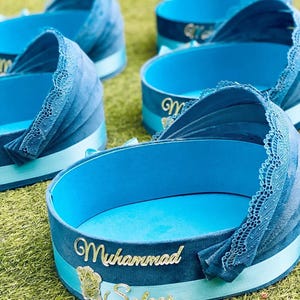 Puede incluir: Varias cunas decorativas azules para bebés con ribete de encaje y detalles de cinta están dispuestas sobre hierba verde. Cada cuna tiene una placa dorada con los nombres "Muhammad" y "Salaar".