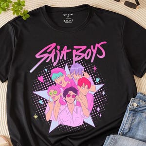 以下が含まれることがあります： ピンクと紫の色合いの5人の漫画のキャラクターのグラフィックと、ピンク色の「Saja Boys」のテキストが特徴の黒いTシャツ。デザインには星とハートが含まれています。