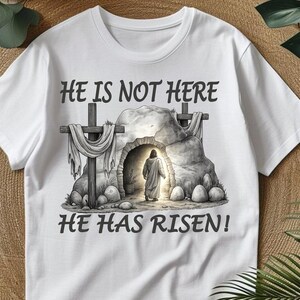 May include: White t-shirt with the text "HE IS NOT HERE" above an illustration of a figure exiting a tomb, and the text "HE HAS RISEN!" below. The graphic is in shades of gray and brown.