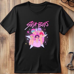 Puede incluir: Camiseta negra con un gráfico de seis personajes de dibujos animados en rosa, morado y verde, y el texto "Saja Boys" en rosa. Las figuras están dispuestas frente a una estrella blanca con un fondo punteado.