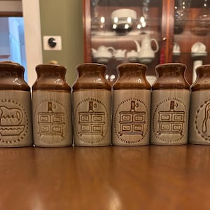 May include: Six vintage ceramic jars with brown lids and tan bodies. Each jar features a different embossed design, including a pitcher, a dresser, and a water jug. The jars are arranged in a row on a wooden surface.