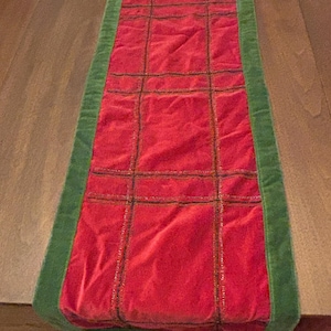 May include: A rectangular red table runner with a green border. The center of the runner is divided into rectangular sections with dark stitching. The runner is made of a soft, velvety material.