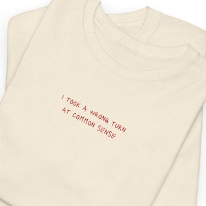Op de afbeelding: Twee crèmekleurige t-shirts zijn op elkaar gestapeld. Het bovenste shirt heeft de geborduurde tekst "I took a wrong turn at common sense" in rode draad. De shirts zijn netjes opgevouwen, met de halslijn zichtbaar.