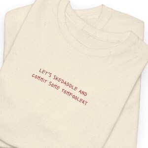Può includere: Due magliette color crema sono impilate. La maglietta superiore ha la frase "LET'S SKEDADDLE AND COMMIT SOME TOMFOOLERY" in testo rosso. Le magliette sono piegate e sembrano essere fatte di un materiale morbido.
