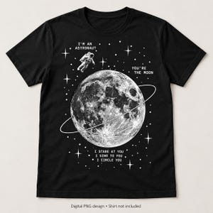 Könnte beinhalten: Ein schwarzes Kurzarm-T-Shirt mit einer weißen Grafik des Mondes mit Ring, einem Astronauten und Sternen. Der Text lautet: I'M AN ASTRONAUT, YOU'RE THE MOON, I STARE AT YOU, I SING TO YOU, I CIRCLE YOU. Das Design ist mittig platziert.