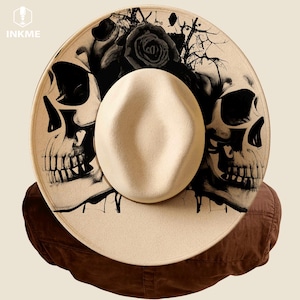 May include: A cream-colored fedora hat with a wide brim, featuring a black skull and rose design. The hat has a structured crown and a contrasting band. The image includes the text "INKME" in the upper left corner.