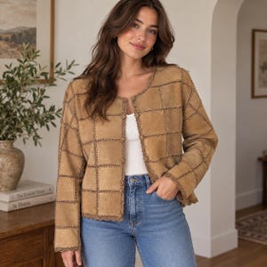 May include: A tan suede jacket featuring a grid-like patchwork design with visible whipstitch detailing along the seams. The jacket has a round neckline, long sleeves, and a front button closure, worn over a plain white top and blue jeans.