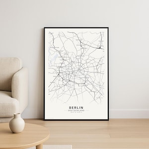 Könnte beinhalten: Eine gerahmte schwarz-weiße Karte von Berlin, Deutschland. Die Karte zeigt das Straßennetz der Stadt und ist mit "Berlin Deutschland" und Koordinaten beschriftet. Das Kunstwerk wird an einer weißen Wand mit einem beigen Sofa und einem Holztisch im Vordergrund gezeigt.