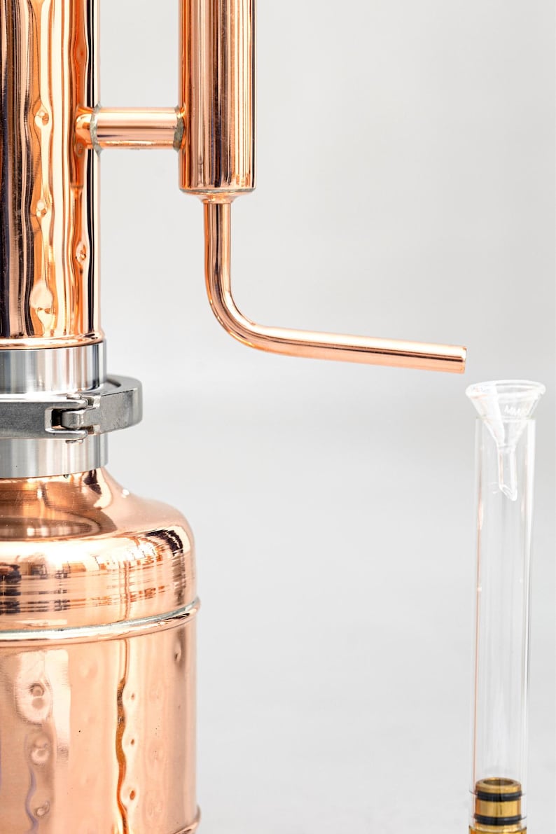 Pu&ograve; includere: Un alambicco in rame con un beccuccio curvo e una provetta di vetro trasparente. L'alambicco ha una superficie lucida e riflettente ed &egrave; progettato per la distillazione. L'immagine &egrave; su sfondo bianco.