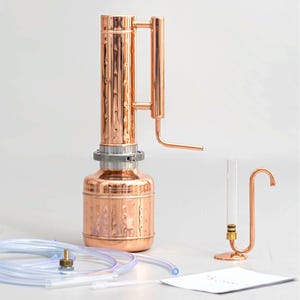 Pu&ograve; includere: Un alambicco in rame con una colonna cilindrica e una base rotonda, con un condensatore e un tubo di vetro trasparente. Tubi trasparenti e un piccolo raccordo in rame sono in primo piano. &Egrave; presente anche un piccolo supporto in rame con un tubo curvo.