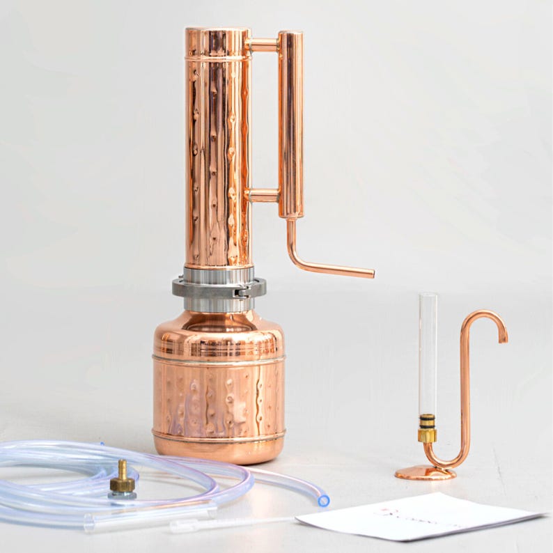 Pu&ograve; includere: Un alambicco in rame con una colonna cilindrica e una base rotonda, con un condensatore e un tubo di vetro trasparente. Tubi trasparenti e un piccolo raccordo in rame sono in primo piano. &Egrave; presente anche un piccolo supporto in rame con un tubo curvo.