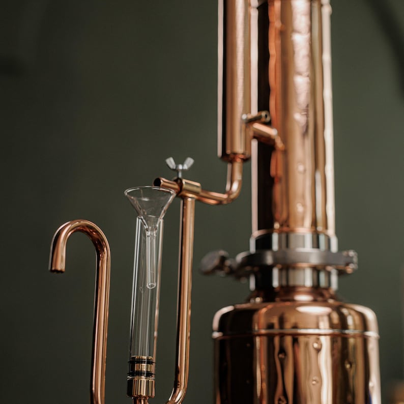 Pu&ograve; includere: Primo piano di un alambicco in rame con un imbuto di vetro e un beccuccio curvo. L'alambicco ha un corpo cilindrico e vari tubi e raccordi. Lo sfondo &egrave; di colore verde scuro.