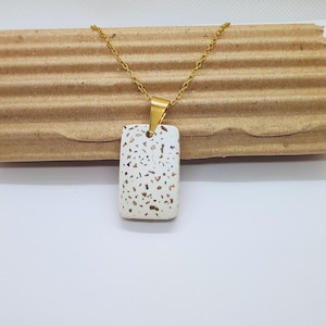 Pode incluir: Um colar com pingente retangular e corrente dourada. O pingente é branco com pequenas manchas marrons irregulares. O pingente é suspenso por uma fiança dourada. O colar é exibido em uma superfície de papelão ondulado.