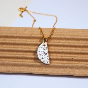 Pode incluir: Colar com corrente dourada e pingente. O pingente é branco, em forma de meia-lua, com pequenas manchas coloridas. O colar é exibido em uma superfície de papelão ondulado.