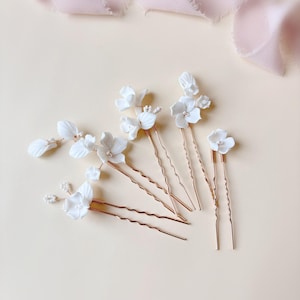 Puede incluir: Un conjunto de horquillas para el cabello con flores blancas y tallos color cobre. Las horquillas presentan delicados diseños florales tridimensionales, perfectos para peinados de novia o de ocasiones especiales. Las horquillas están dispuestas sobre una superficie beige claro con una cinta rosa en el fondo.