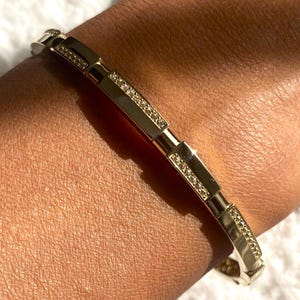 May include: A gold bracelet with a rectangular design and small, sparkling embellishments. The bracelet is worn on a wrist, showcasing its elegant and detailed craftsmanship. The jewellery piece is set against a soft, white background.