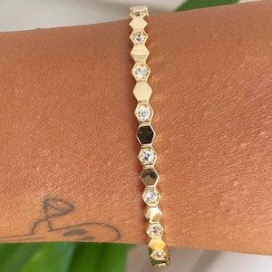 Op de afbeelding: Een goudkleurige armband met een reeks zeshoekige schakels, afgewisseld met heldere, ronde edelstenen. De armband wordt om een arm gedragen, wat het delicate ontwerp en de glans benadrukt. De sieraden zijn een stijlvol accessoire.