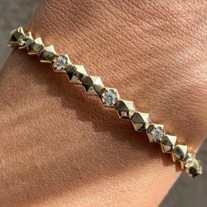 May include: Gold-toned bracelet featuring a geometric design with alternating diamond-like accents. The bracelet is worn on a wrist, showcasing its delicate and elegant appearance. A stylish piece of jewelry.