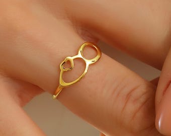 Può includere: Un anello dorato con un design a forma di cuore. L'anello è indossato su un dito, evidenziando il suo stile delicato ed elegante. Il design dell'anello incorpora un cuore e un anello, creando un gioiello unico e accattivante.