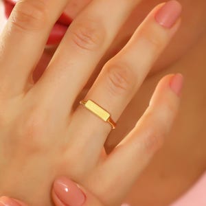 May include: A gold-colored ring with a rectangular design is worn on a finger. The ring is the focal point, set against a background of a pink collared shirt and a person's skin. The ring's simple design is highlighted by the soft lighting.