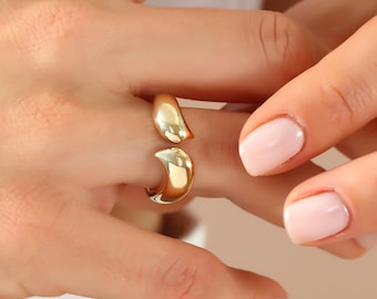 May include: A gold-colored ring with a unique open design. The ring has a smooth, polished surface and a curved shape. The ring is worn on a finger. The background is a white shirt and a neutral background.