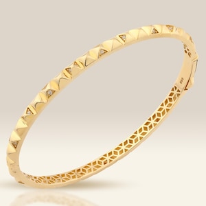 May include: A gold-coloured bangle bracelet with a geometric design. The bracelet features pyramid-shaped accents along the top and a patterned design on the inside. The jewellery is displayed on a light background.