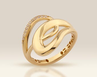 May include: A gold-colored ring with an abstract design. One band is adorned with small, sparkling stones. The ring features a flowing, intertwined design with a polished finish. The ring is displayed on a reflective surface.