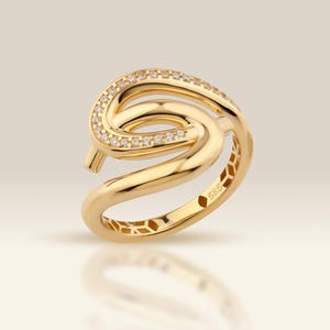 May include: A gold ring with a swirling design, set with small, sparkling stones. The ring has a polished finish and an openwork pattern on the band. The ring is displayed on a reflective surface.